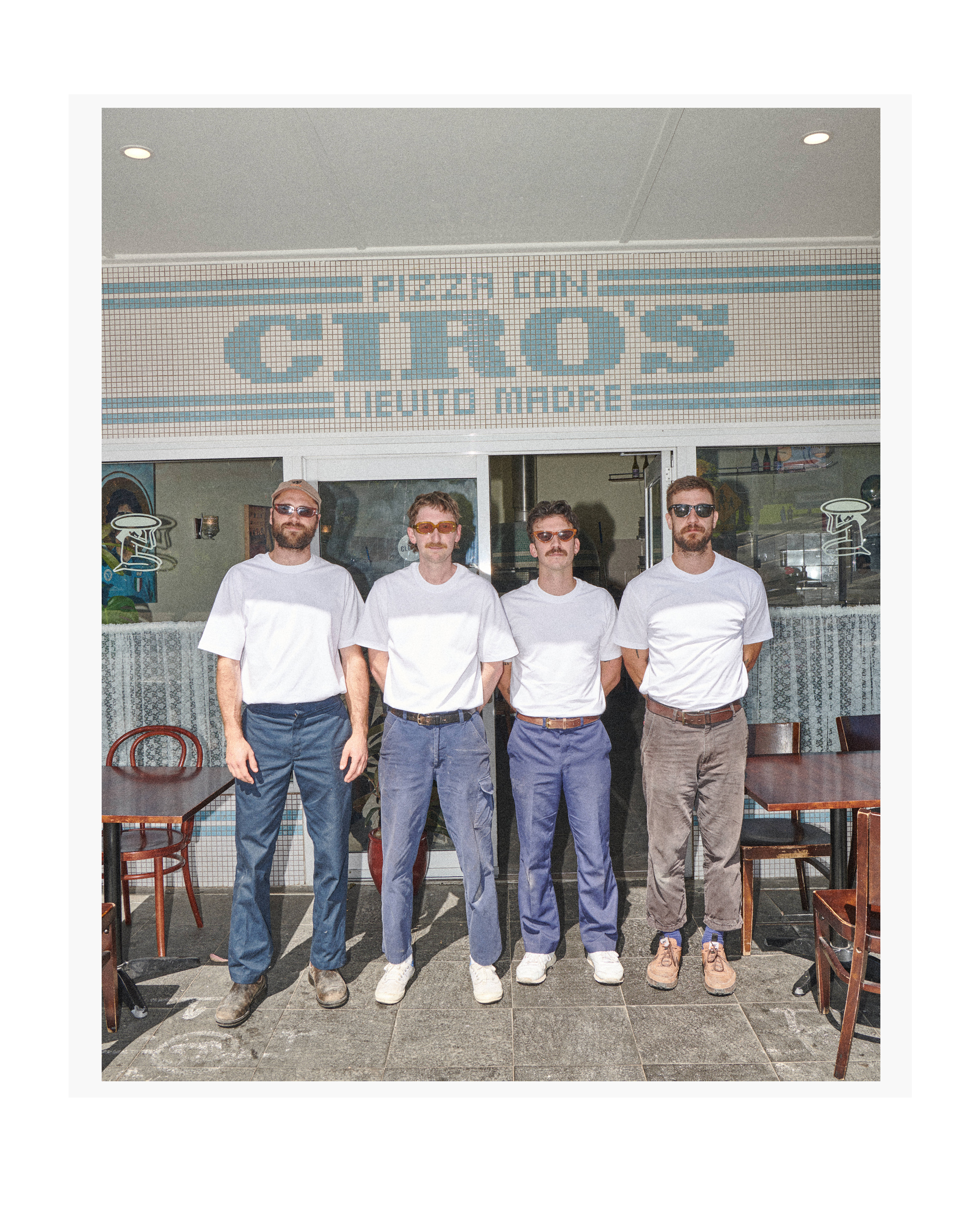 Four men in casual clothing stand outside Ciro's Pizza with a retro aesthetic, captured in a medium shot during daylight.
