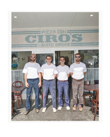 Four men in casual clothing stand outside Ciro's Pizza with a retro aesthetic, captured in a medium shot during daylight.