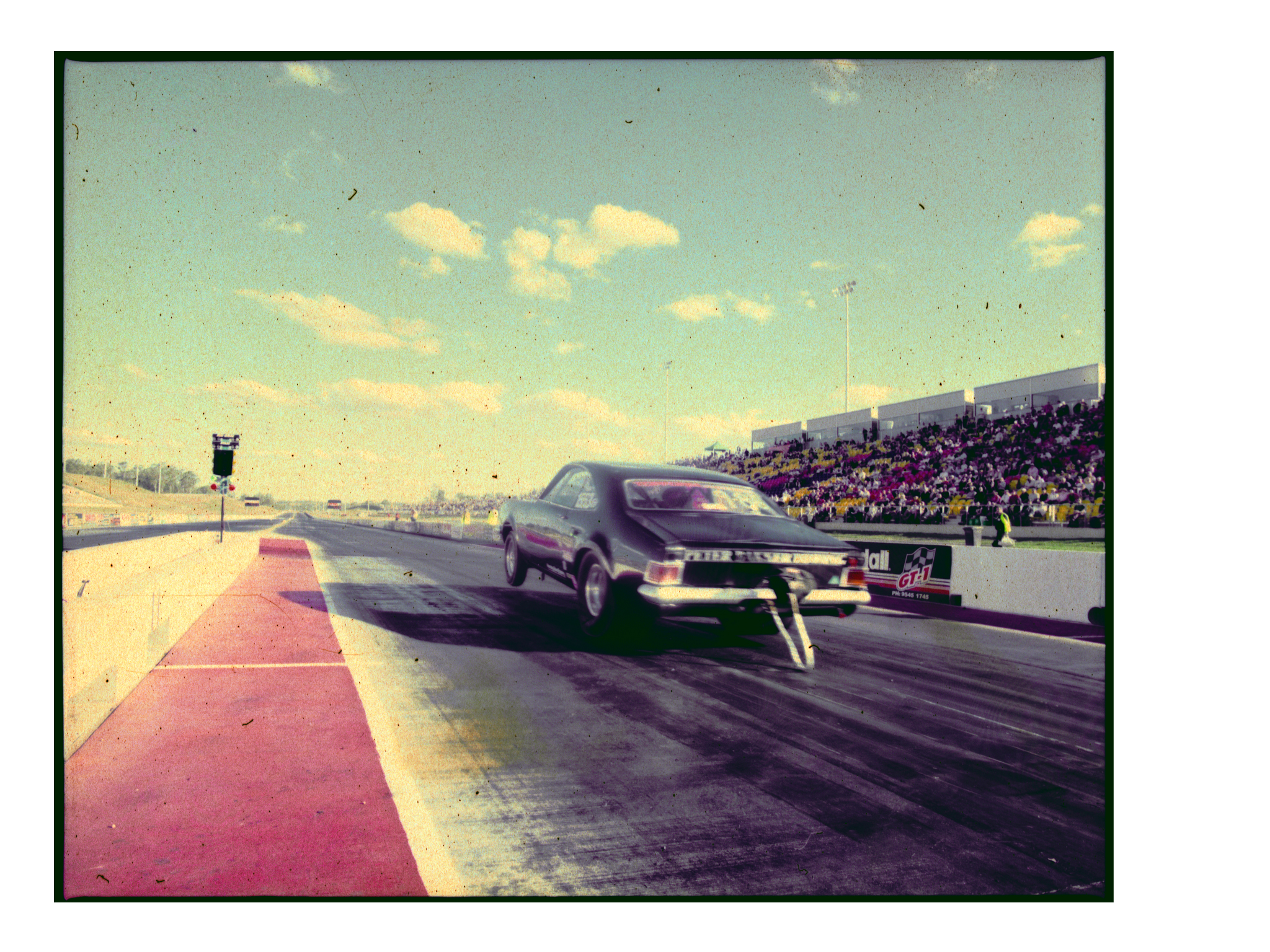 A black drag racing car speeds down the track in an outdoor daytime shot, showing a crowd of spectators in the background.