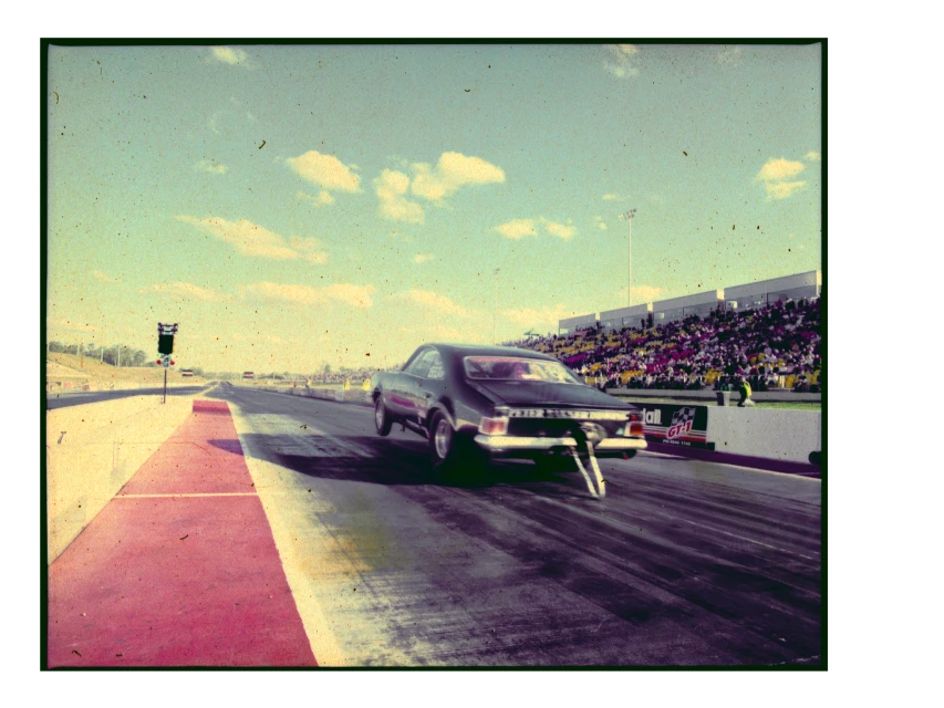 A black drag racing car speeds down the track in an outdoor daytime shot, showing a crowd of spectators in the background.