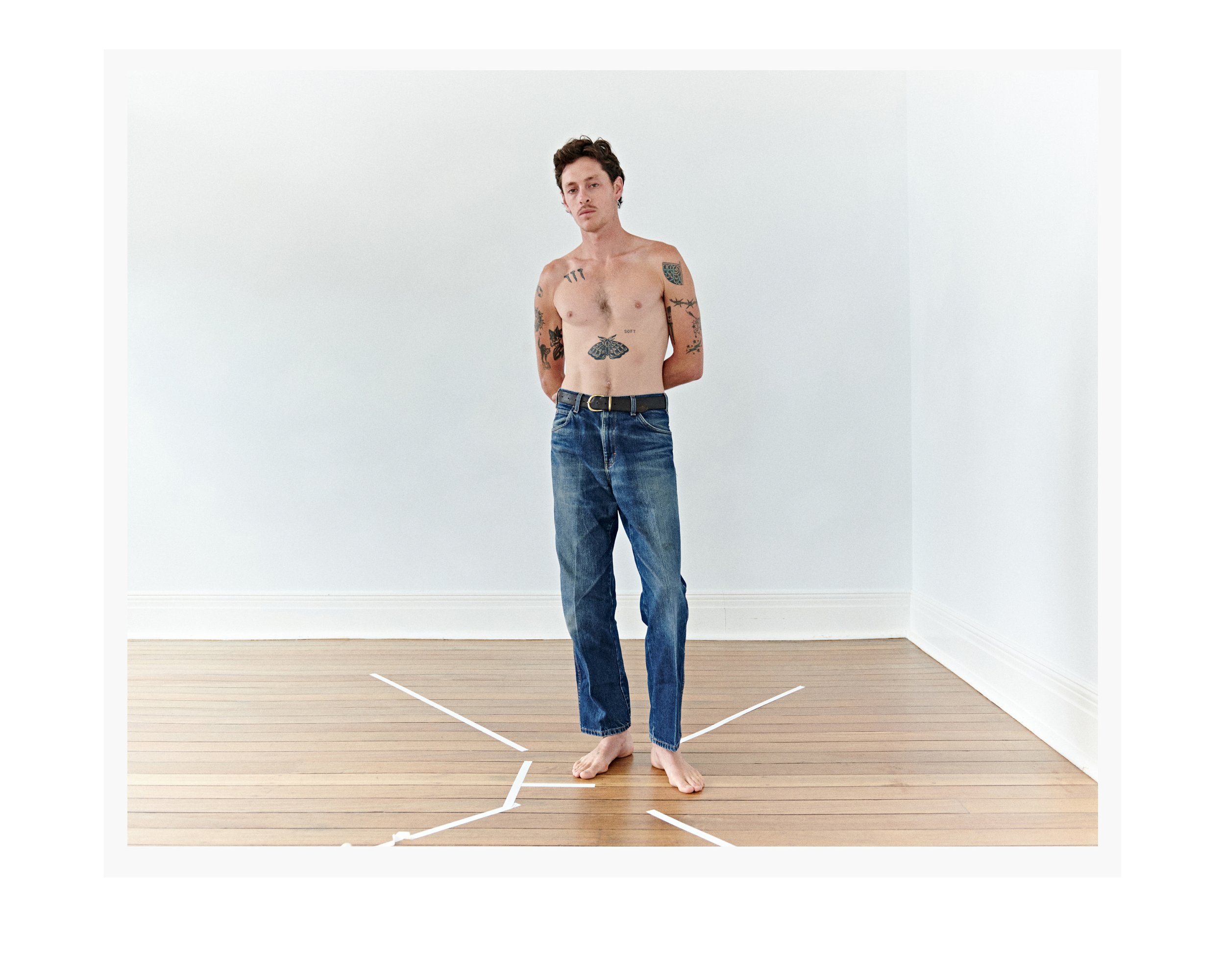 A shirtless man with tattoos standing barefoot in a minimalist white room with a wood floor wearing blue jeans and a black belt.