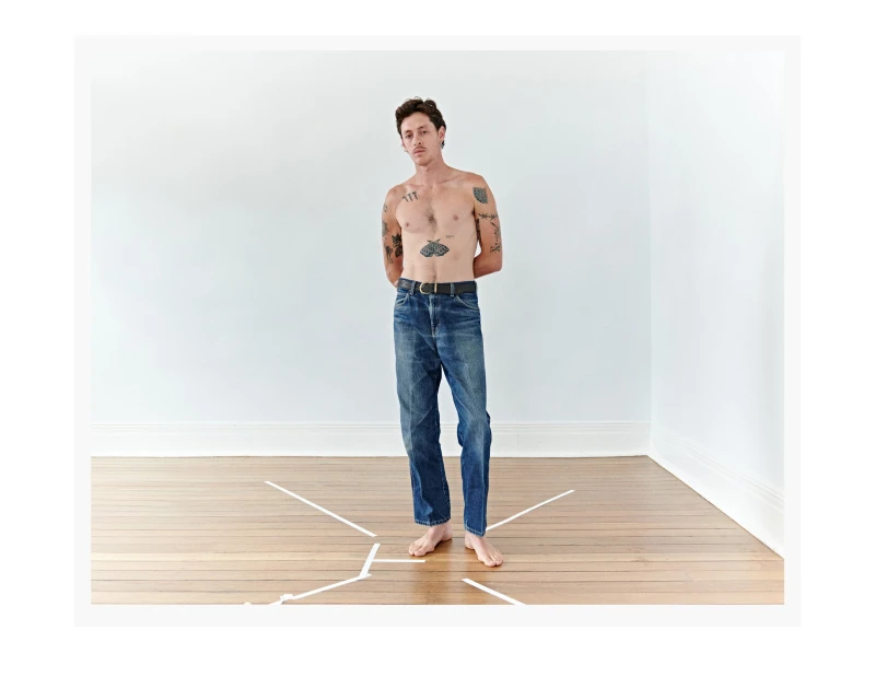 A shirtless man with tattoos standing barefoot in a minimalist white room with a wood floor wearing blue jeans and a black belt.