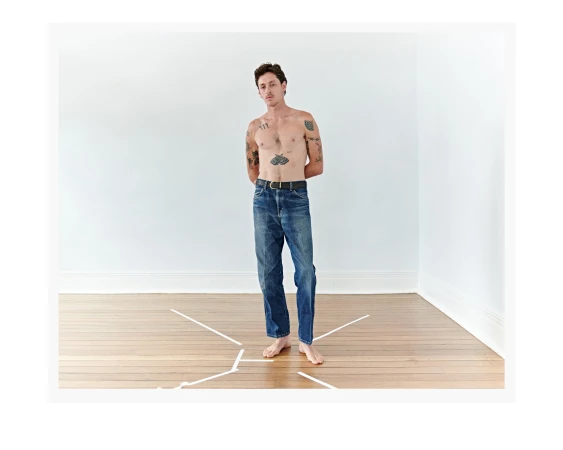 A shirtless man with tattoos standing barefoot in a minimalist white room with a wood floor wearing blue jeans and a black belt.