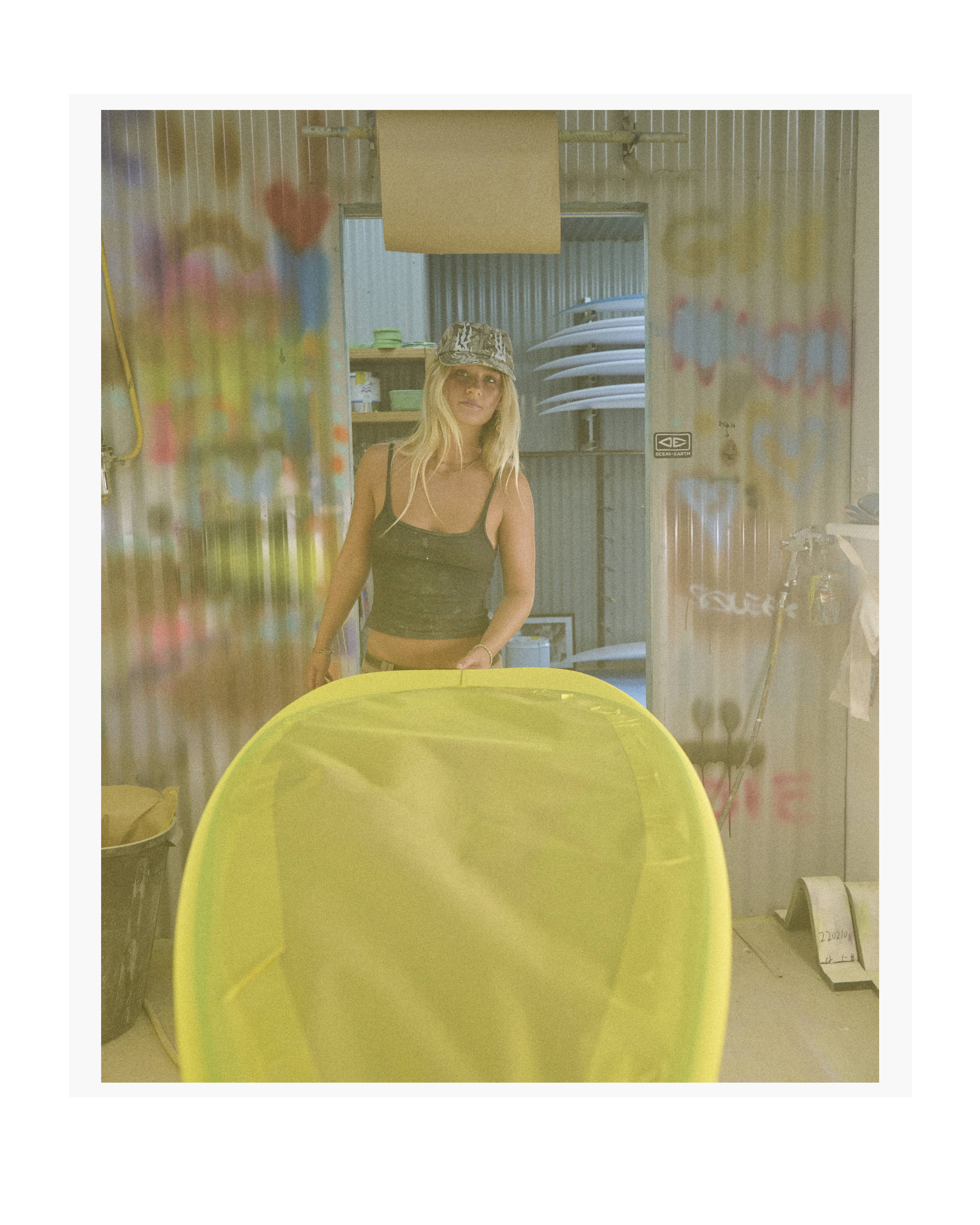 Woman standing in a workshop with surfboards, wearing a camouflage hat and a green tank top. The room has painted corrugated metal walls.