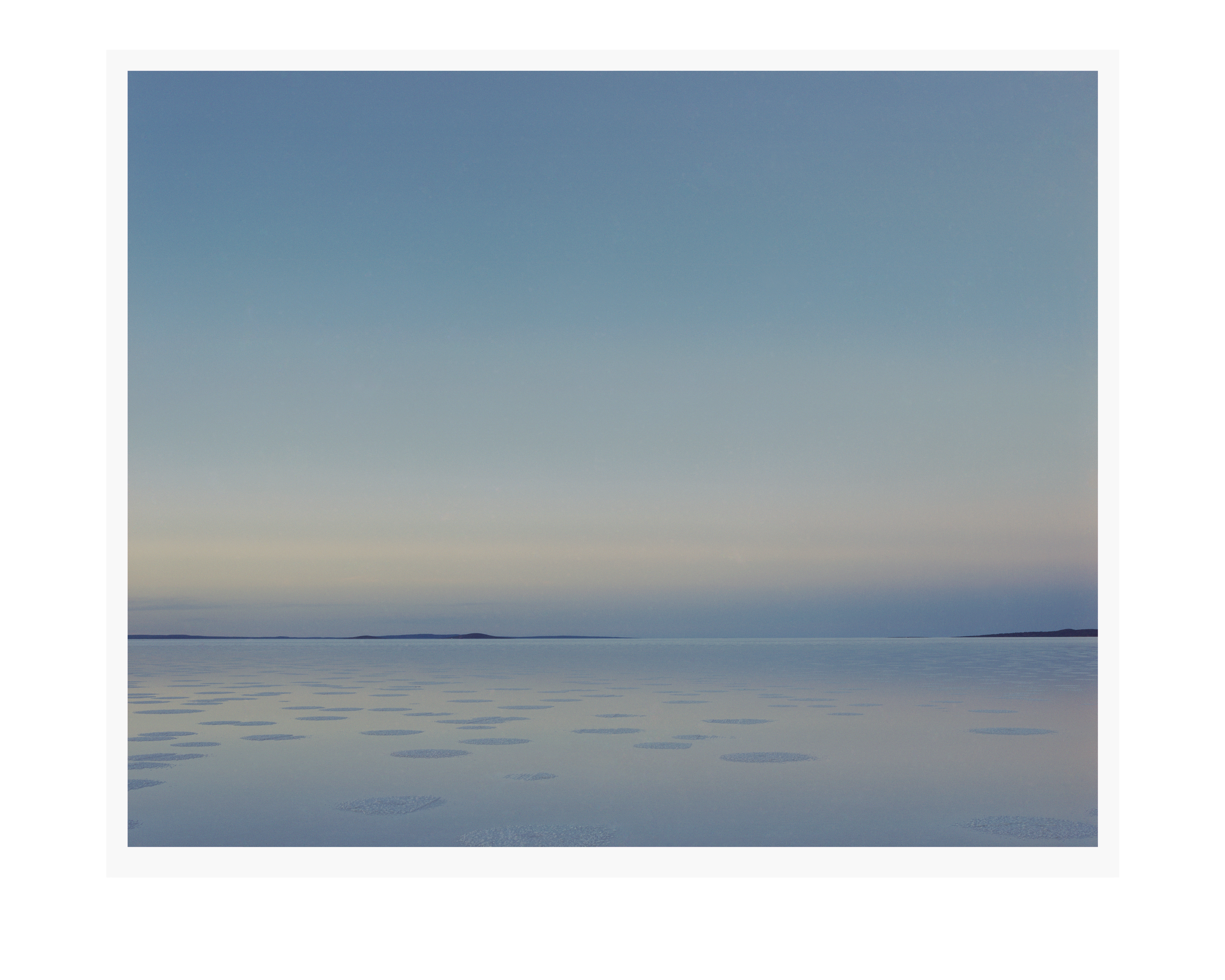 Horizon view of a body of water with scattered ice floes, meeting a light blue sky and distant land, creating a serene landscape.