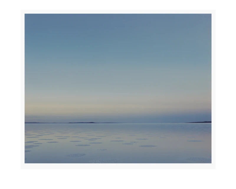 Horizon view of a body of water with scattered ice floes, meeting a light blue sky and distant land, creating a serene landscape.