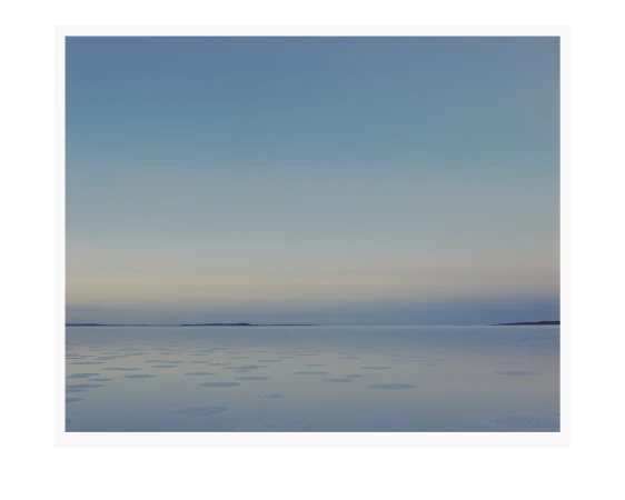 Horizon view of a body of water with scattered ice floes, meeting a light blue sky and distant land, creating a serene landscape.