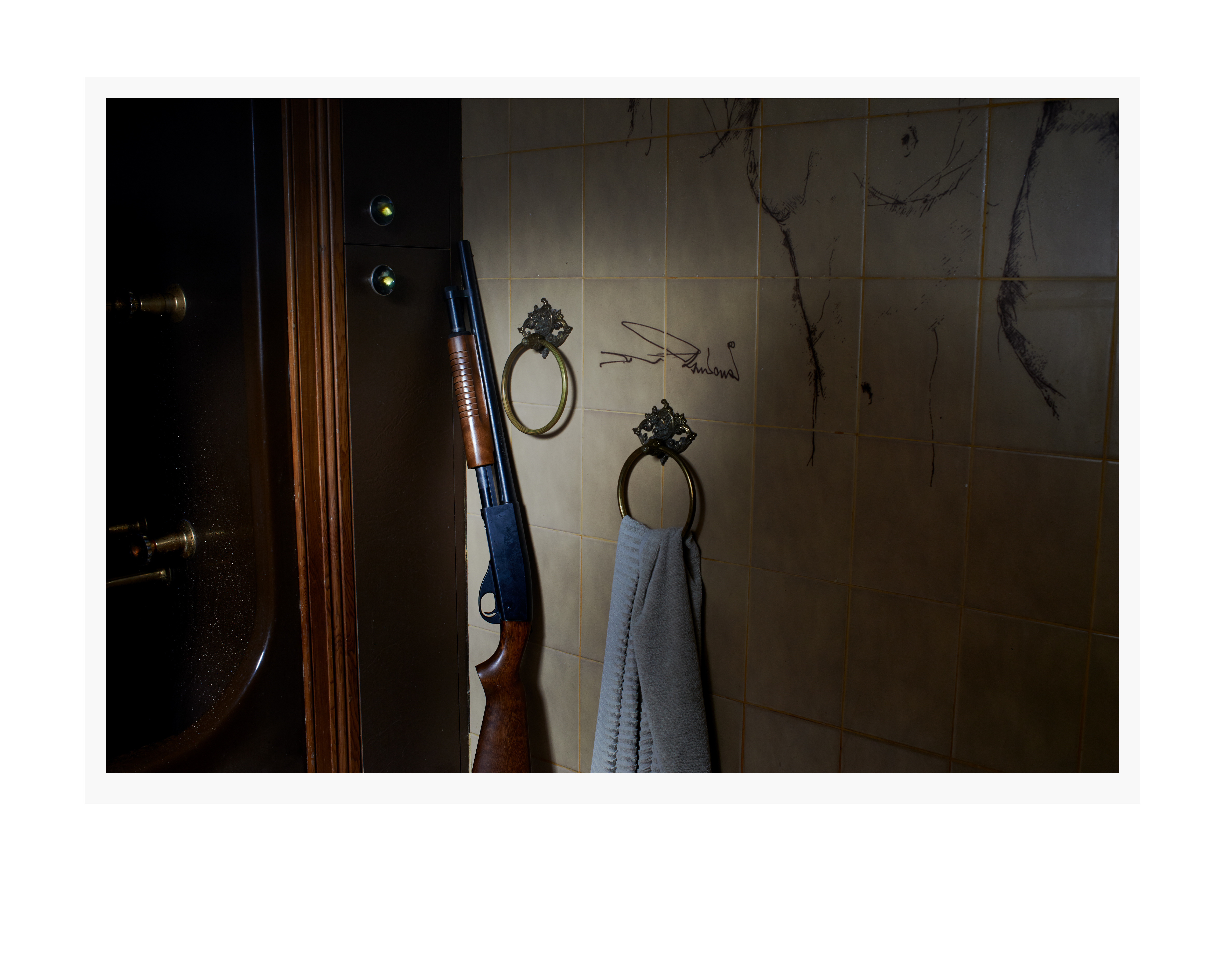 A shotgun leans against a wall in a room with tan tile, near gold towel rings and a dark brown door. Neutral, somber lighting.