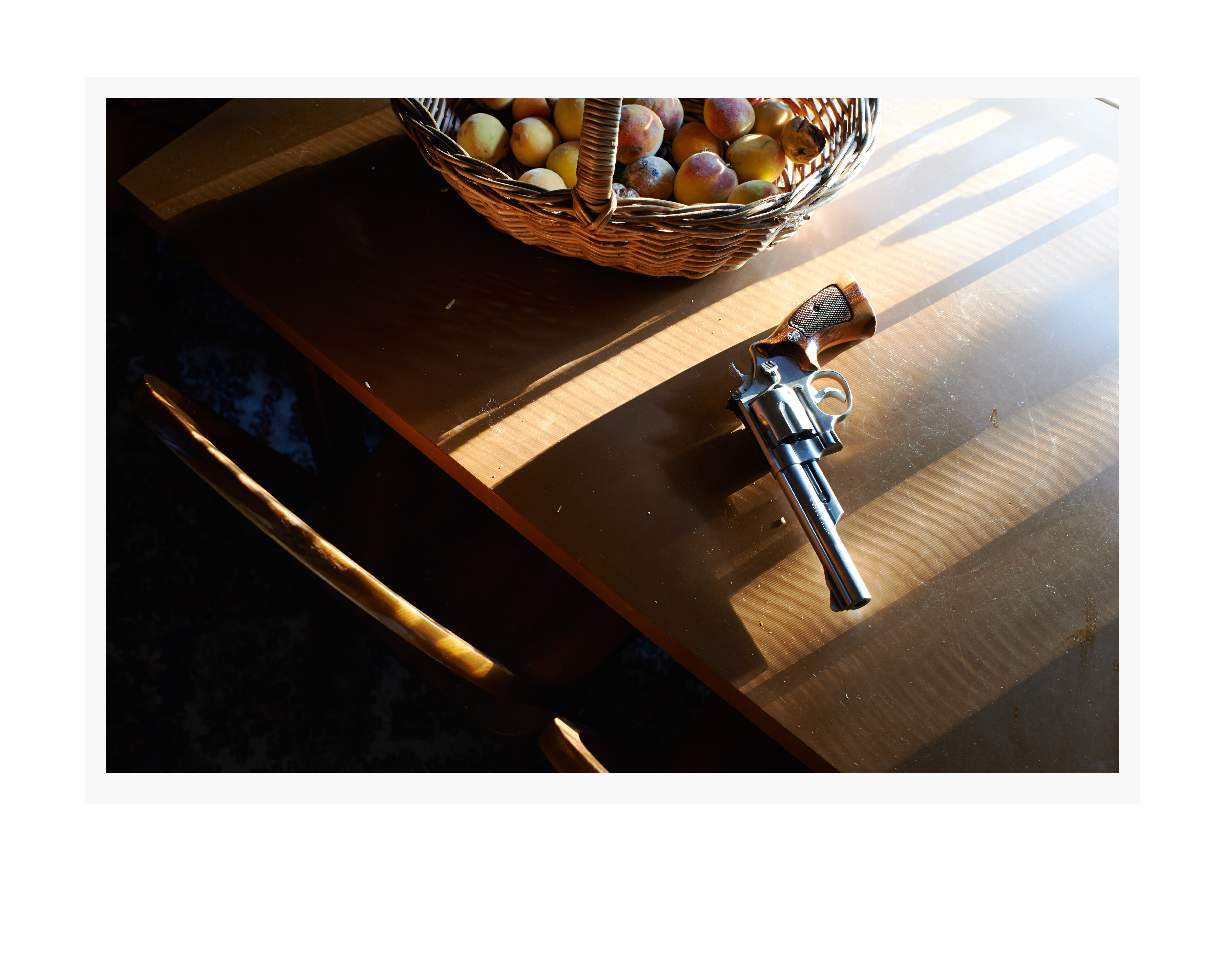 Revolver pistol firearm resting on a wooden table with a basket of fruit, partially illuminated by sunbeams creating shadows.
