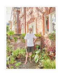 An older man standing barefoot in a courtyard surrounded by potted plants in front of a red brick building.