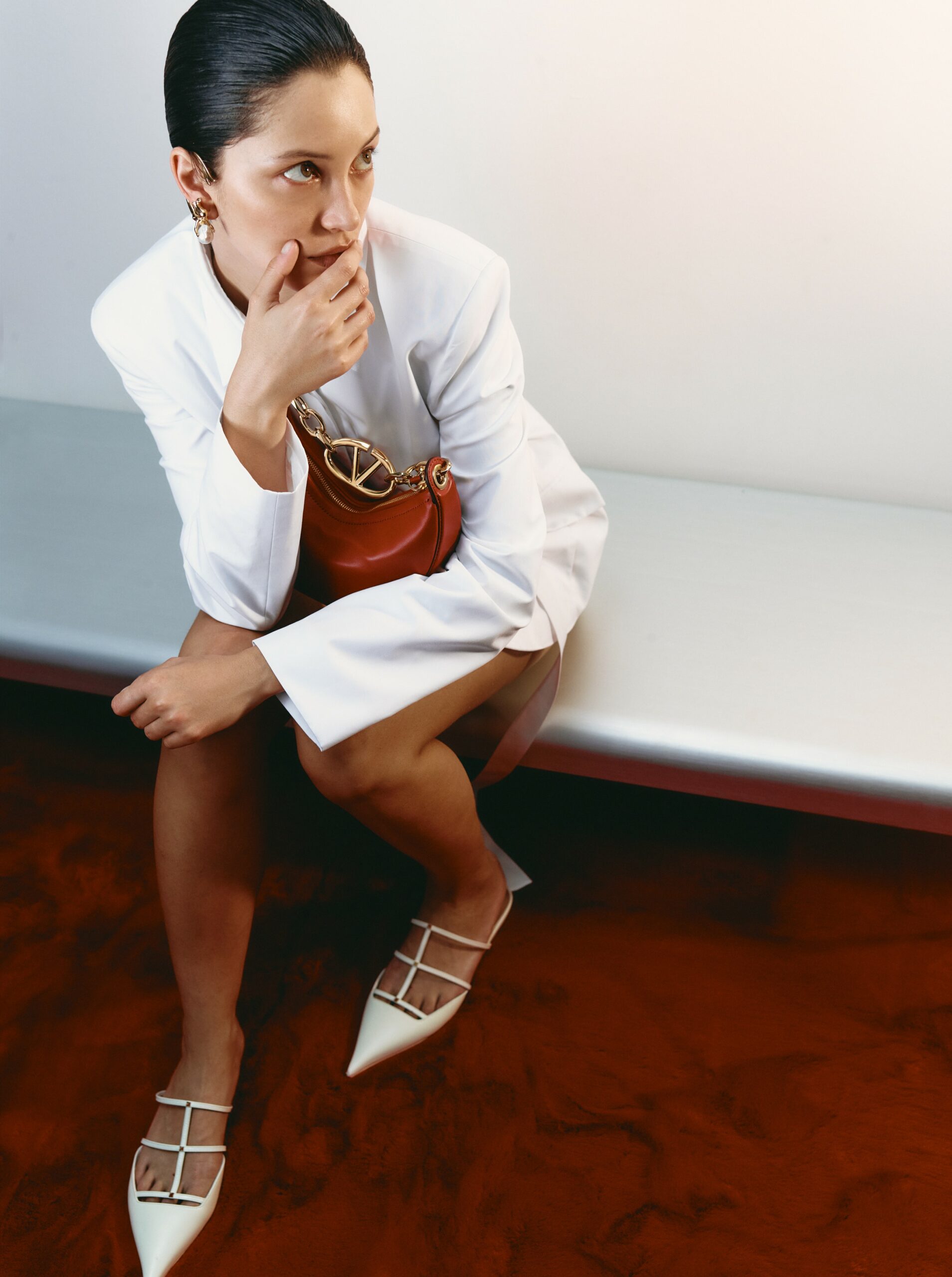 Woman in white suit dress poses on silver bench with red purse and white shoes indoors, gazing off to the side, beauty shot.