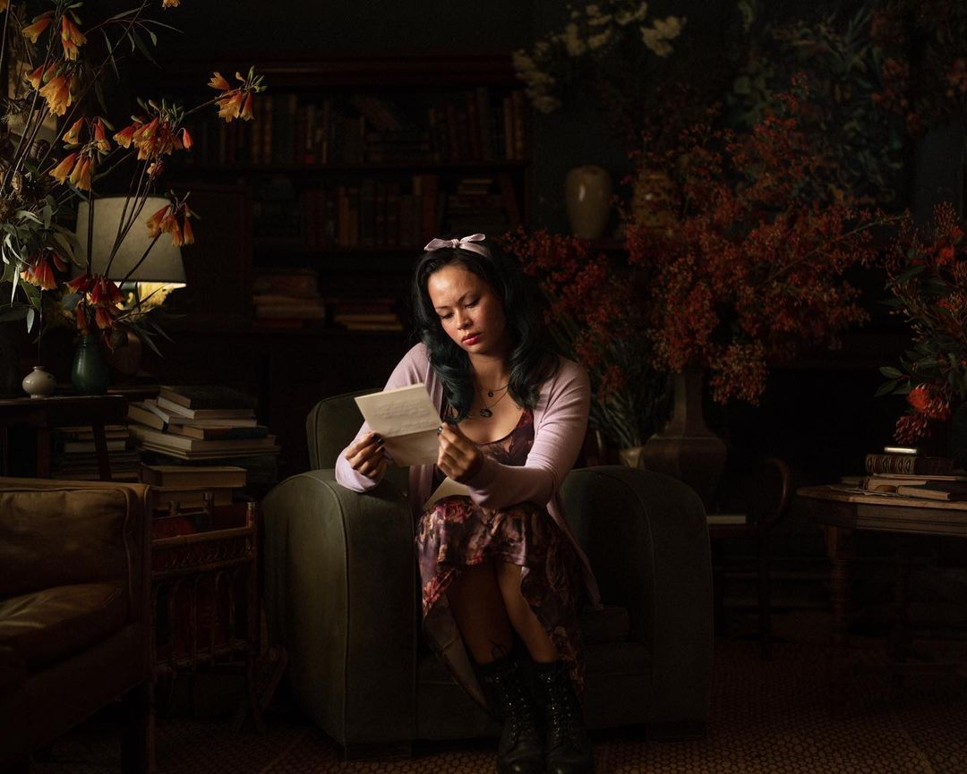 Young woman with a bow in her hair sits in a green armchair and reads a letter indoors surrounded by floral arrangements and books.