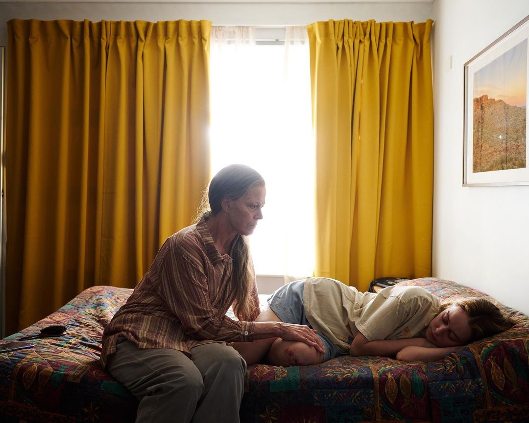 Adult woman sits on bed, touching the leg of a reclining teen girl in an indoor bedroom setting with yellow curtains and neutral tones.