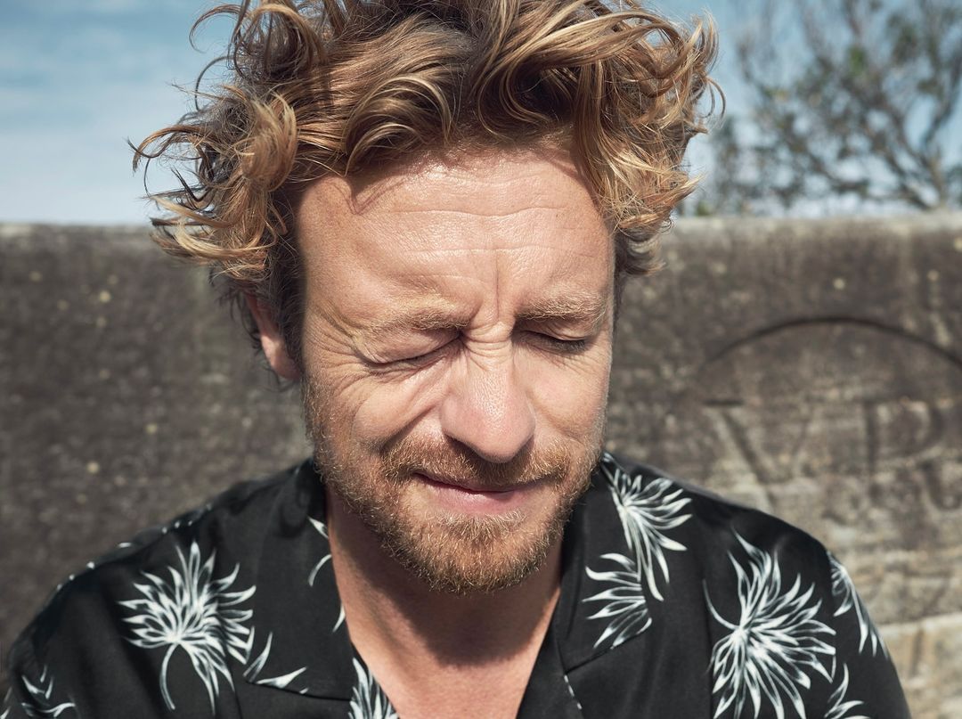 Close-up of a fair-skinned adult male with curly reddish hair, eyes closed, and wearing a floral shirt against a stone backdrop.