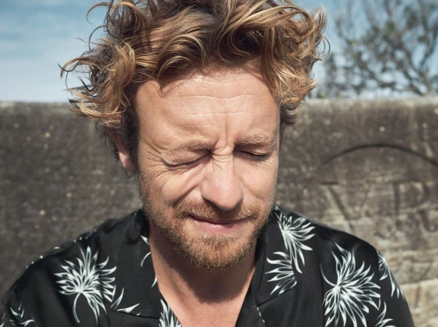 Close-up of a fair-skinned adult male with curly reddish hair, eyes closed, and wearing a floral shirt against a stone backdrop.