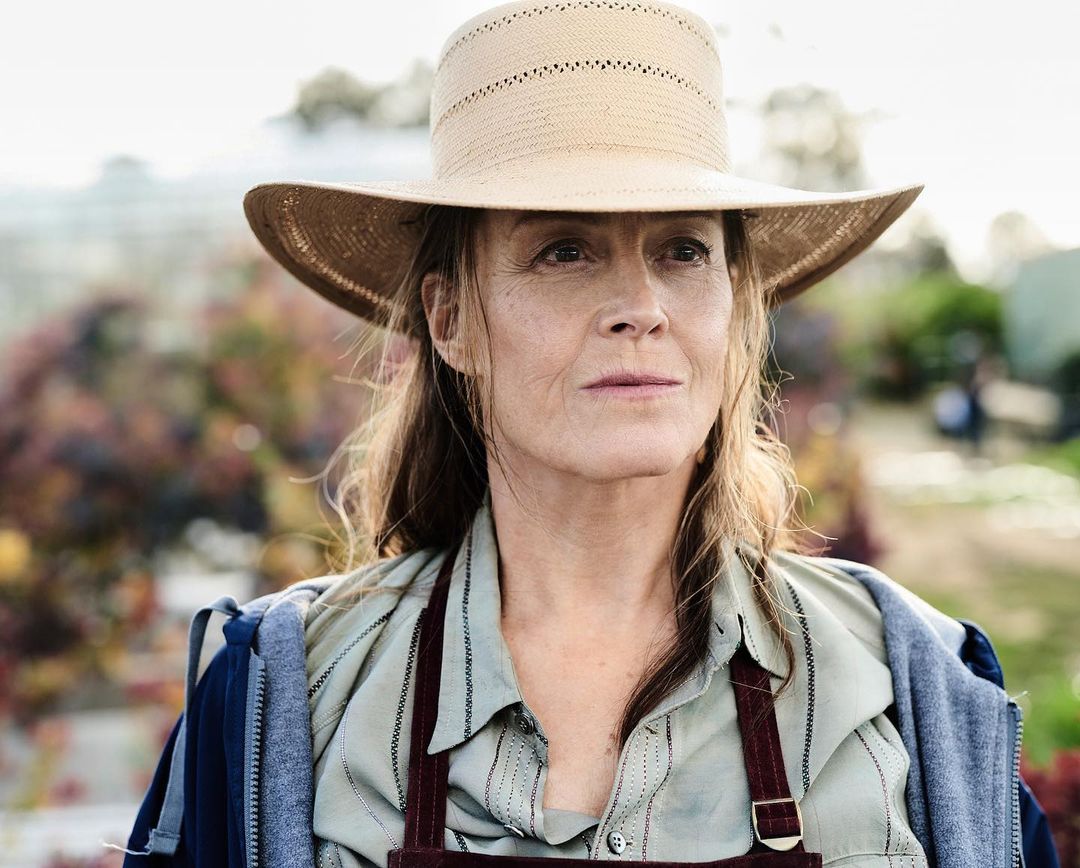 Portrait of a woman with light brown hair wearing a straw hat, striped shirt, and maroon apron, standing in an outdoor garden setting.