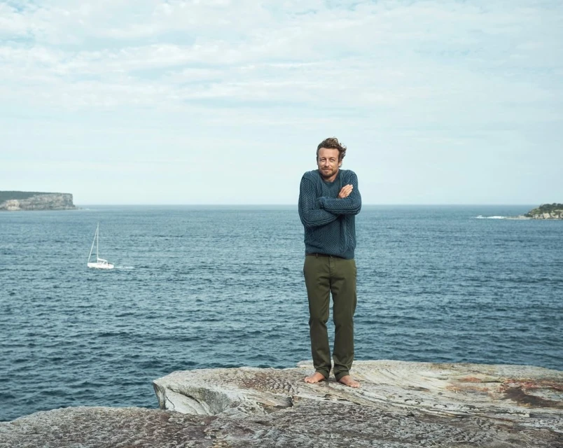 Barefoot man in a blue sweater and green pants stands on a rocky cliff overlooking the ocean with a sailboat in the distance.