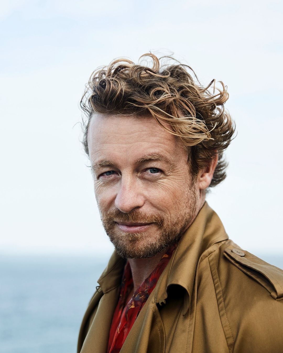Close-up portrait of an adult male with curly hair wearing a trench coat, gazing at the camera against a blurred sky and ocean backdrop.
