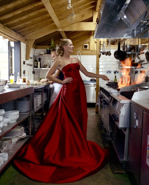 Adult woman in a red evening gown cooks over a gas stove in a commercial kitchen with open flame while an assistant stands nearby.