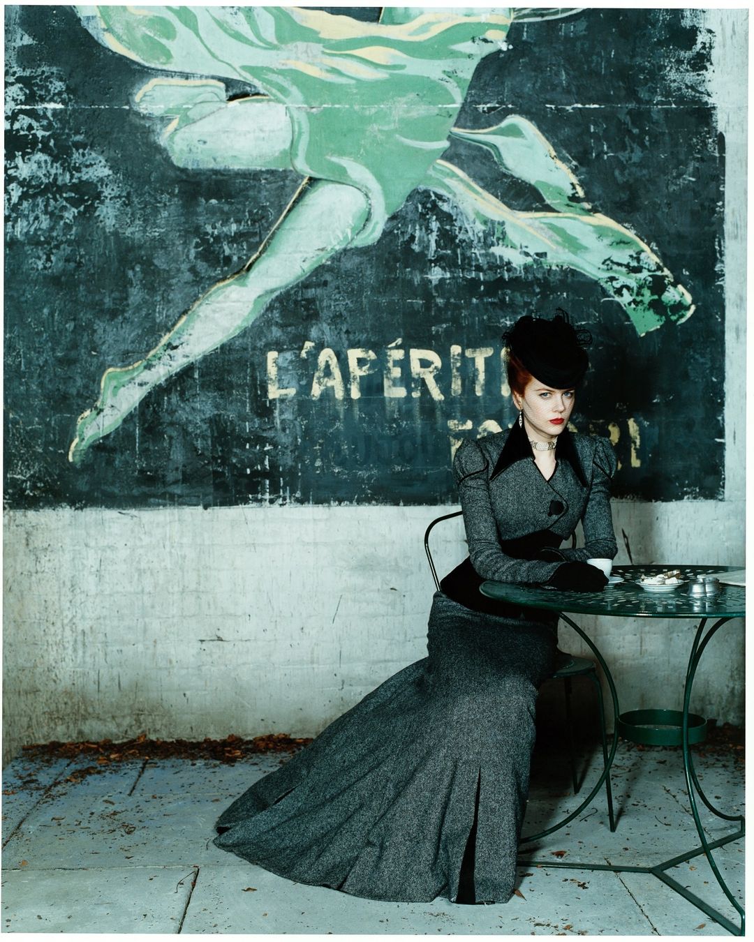 Woman in elegant gray suit and black hat sits at a green metal table in front of a worn advertisement on an outdoor patio.