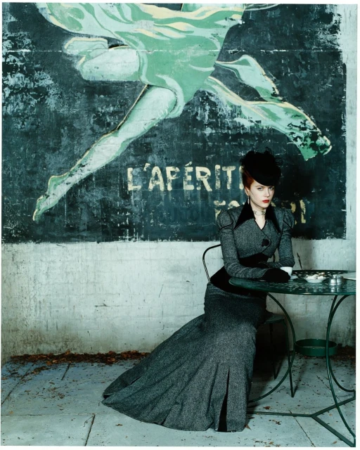 Woman in elegant gray suit and black hat sits at a green metal table in front of a worn advertisement on an outdoor patio.