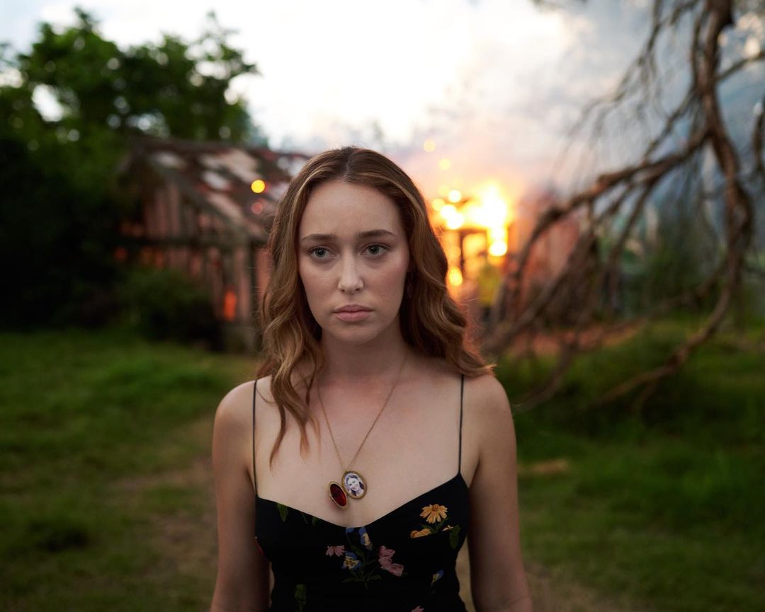 Portrait of a light-skinned woman with long brown hair standing outside a burning building, wearing a floral dress and pendant necklace.