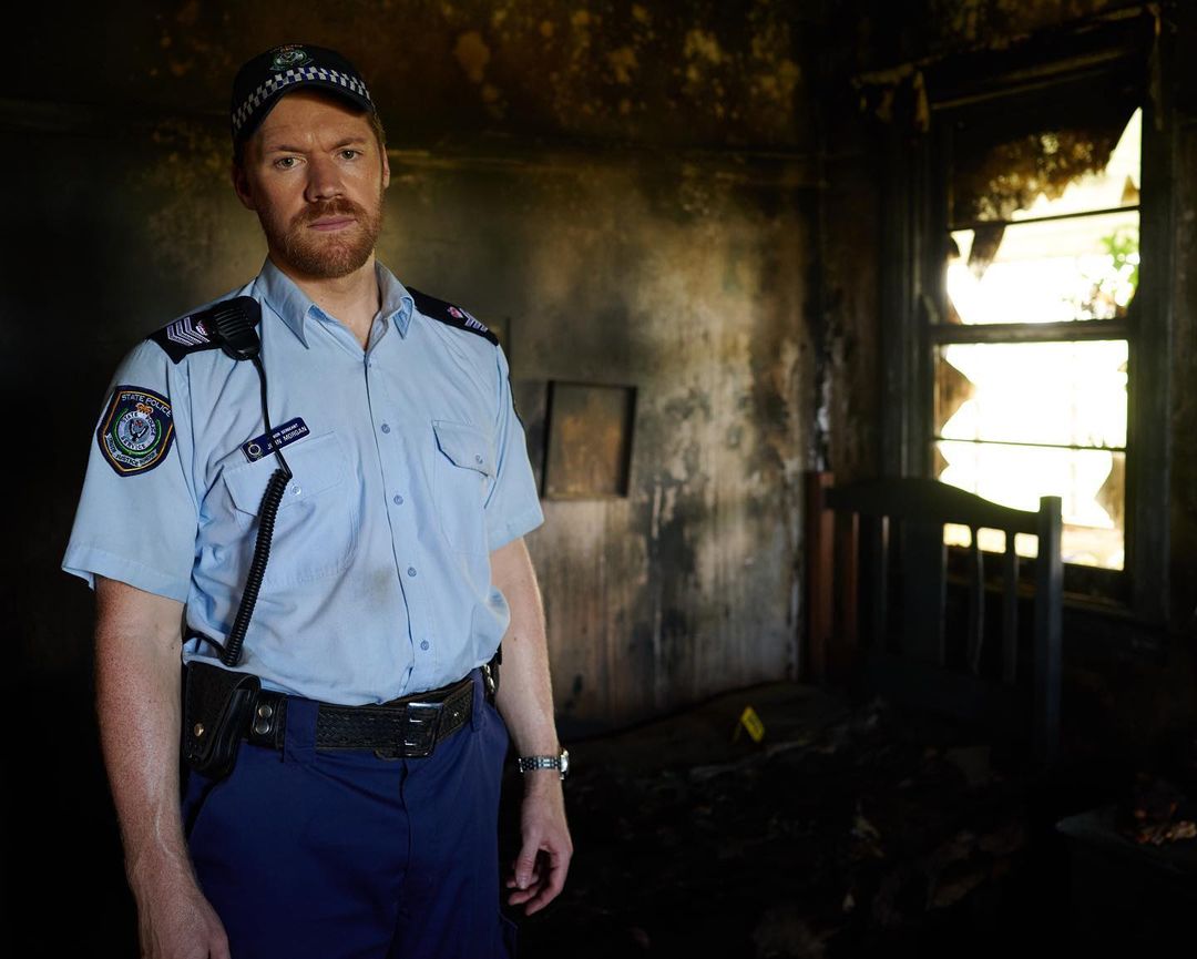 Adult male police officer in uniform stands in a burned room with charred walls and broken window, creating a somber scene.