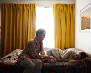 An older woman sits on a patterned bed touching a young woman who lies resting against a window with bright yellow curtains.