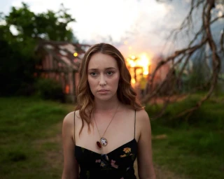Woman in a black floral dress with a locket necklace standing outdoors with a burning building in the background.