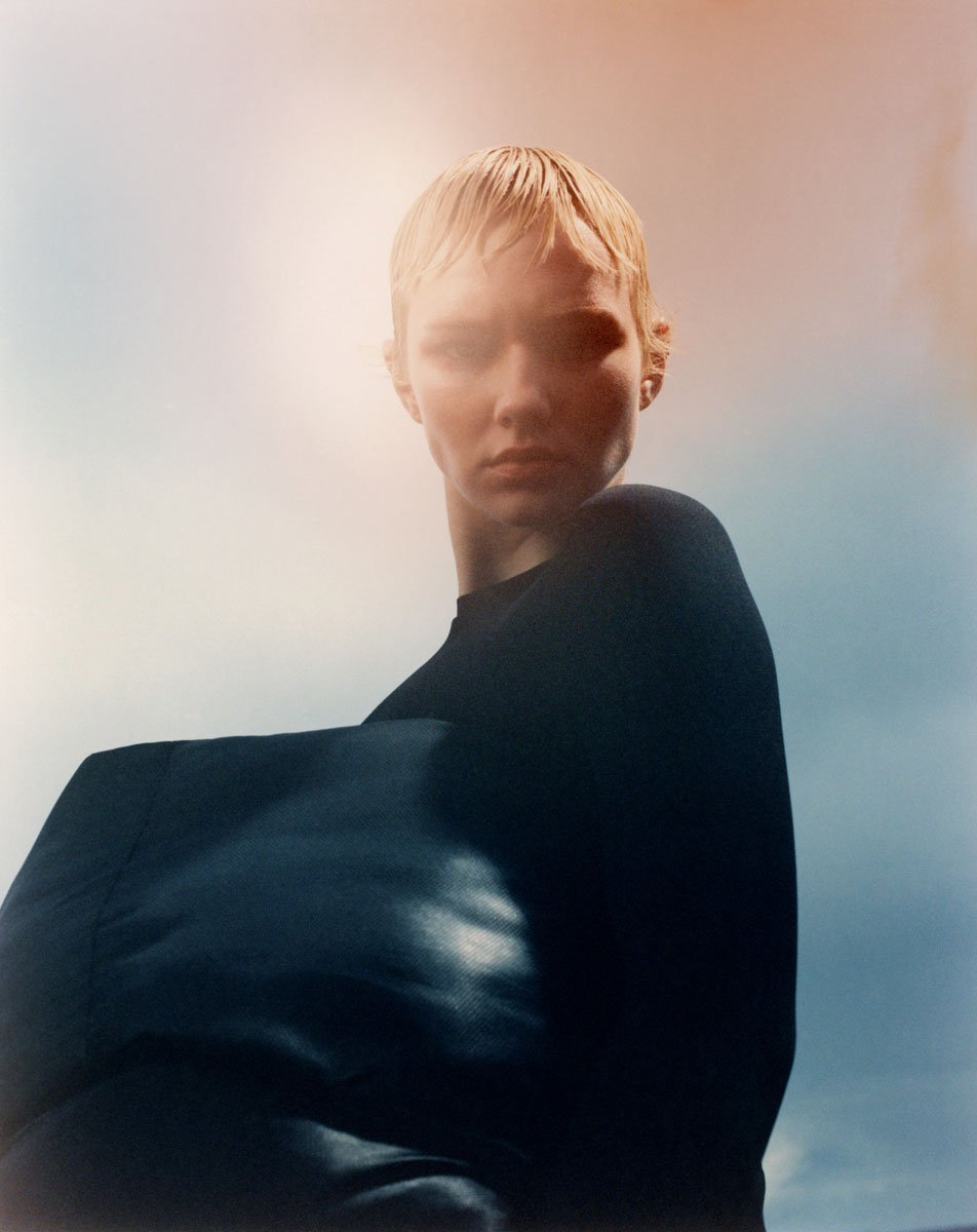 Woman in black long-sleeve shirt poses with soft, diffused lighting and a gradient sky backdrop in a fashion editorial style.