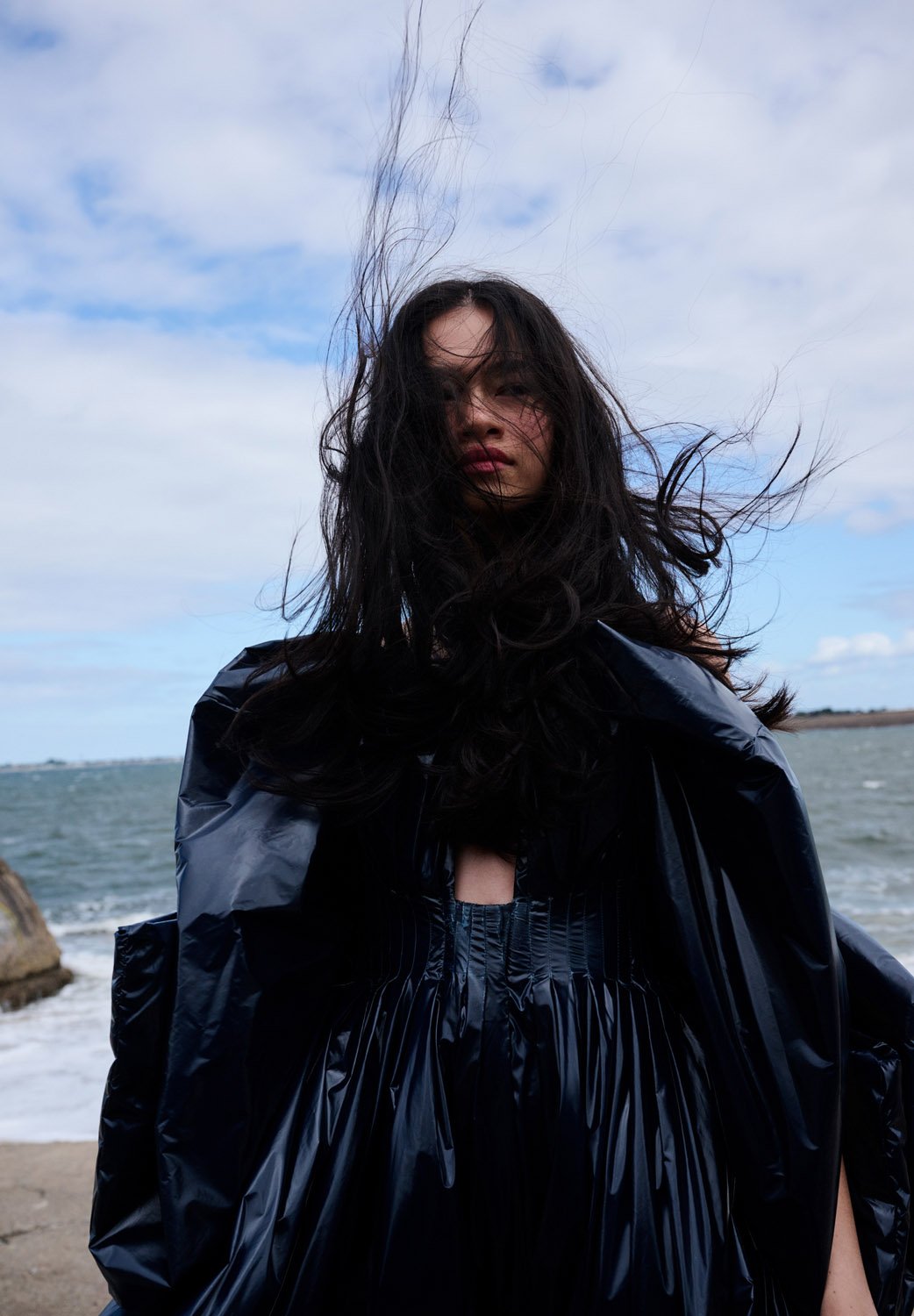 A woman poses on a rocky beach in a shiny black dress with the wind blowing through her hair against a blue sky with clouds.