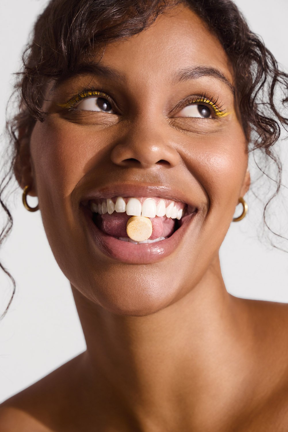 Close-up shot of a smiling young woman with dark curly hair and tanned skin holding a small, round tablet on her tongue indoors.