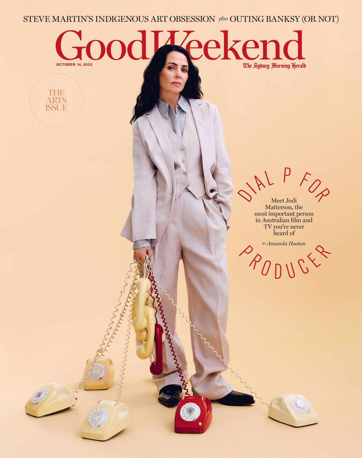 Magazine cover featuring a woman in a suit surrounded by vintage telephones, posing indoors against a light beige background. Editorial shot.