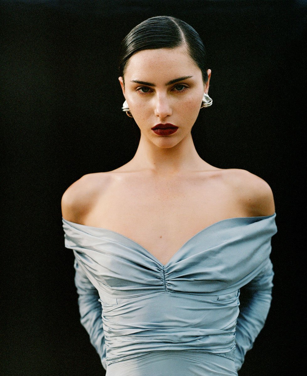 Close-up portrait of a dark-haired woman wearing a light blue dress with large earrings against a dark background in a studio setting.