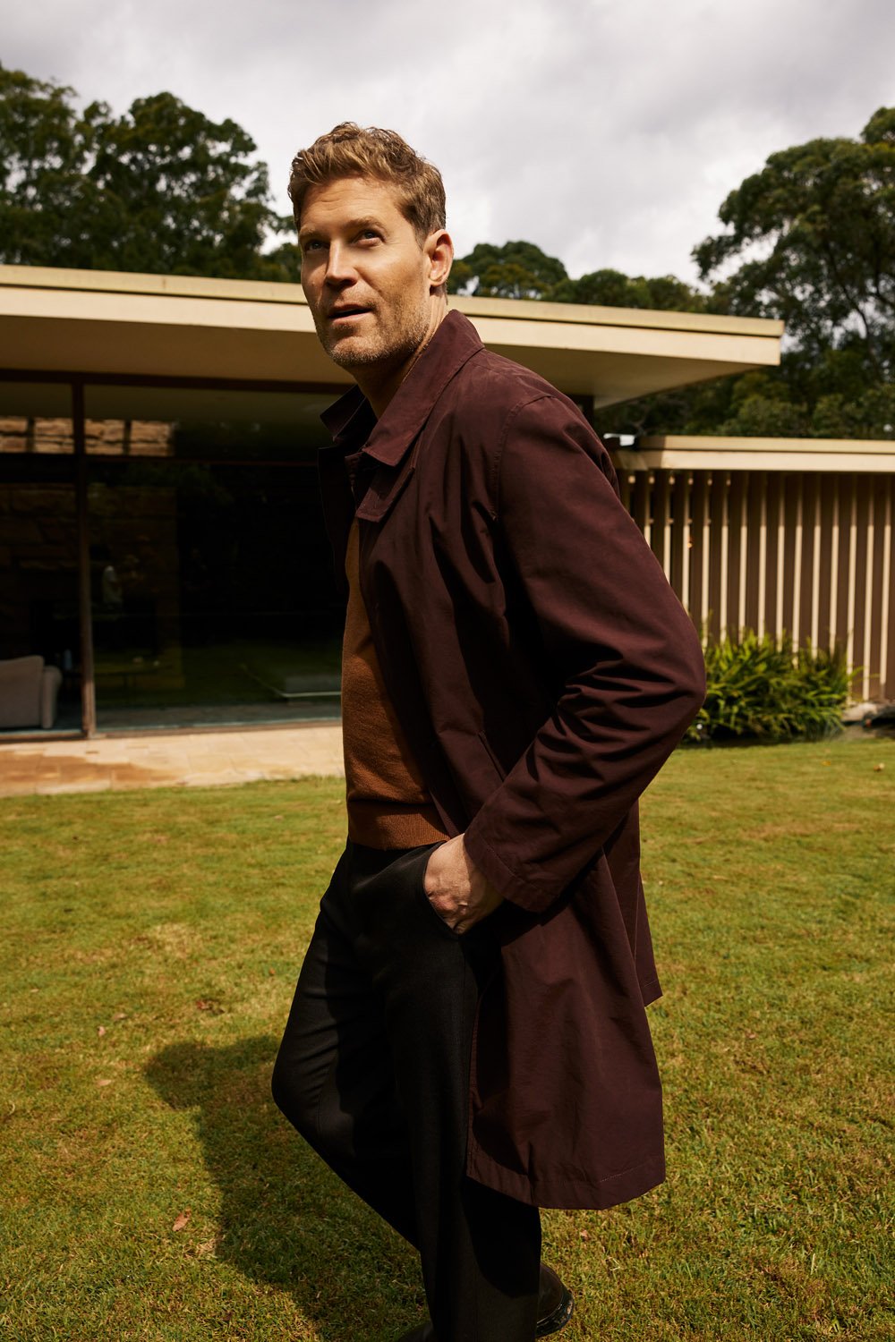 A blond male model poses outdoors on a lawn in a maroon coat and dark pants, looking off-camera with soft daylight.