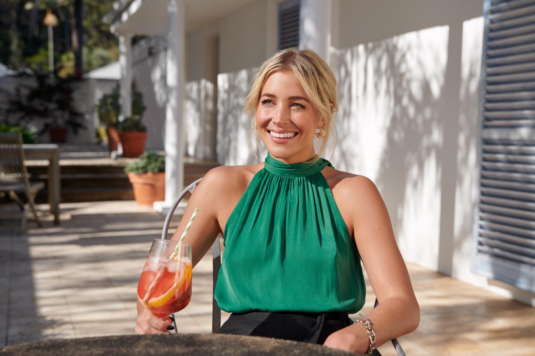 Smiling blonde woman in green top sits outside with cocktail, enjoying sunny day. Louvered doors and greenery create a relaxed backdrop.