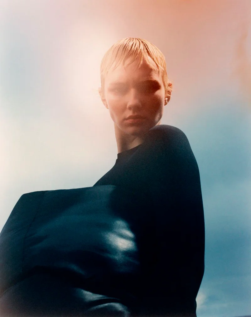 Model with short wet hair and dark top shot from a low angle against a soft sky blue and peach gradient background.