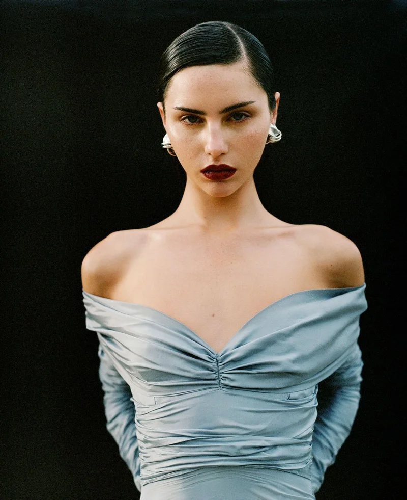 Close-up portrait of a woman with slicked-back dark hair wearing a powder blue off-shoulder top and sculptural silver earrings against black