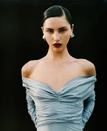 Close-up portrait of a woman with slicked-back dark hair wearing a powder blue off-shoulder top and sculptural silver earrings against black