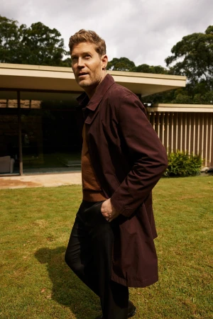 Man with brown hair wearing a burgundy coat standing outdoors on a green lawn in front of a modern house in Sydney.