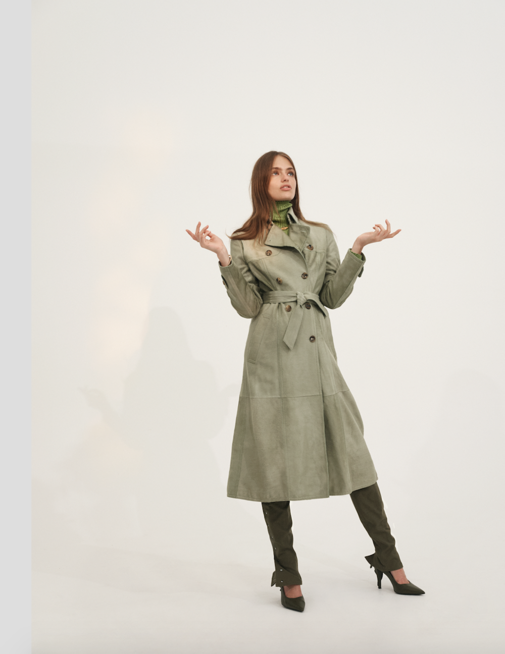 Full-length shot of a woman in a green trench coat and matching boots standing in a white photo studio with arms outstretched.