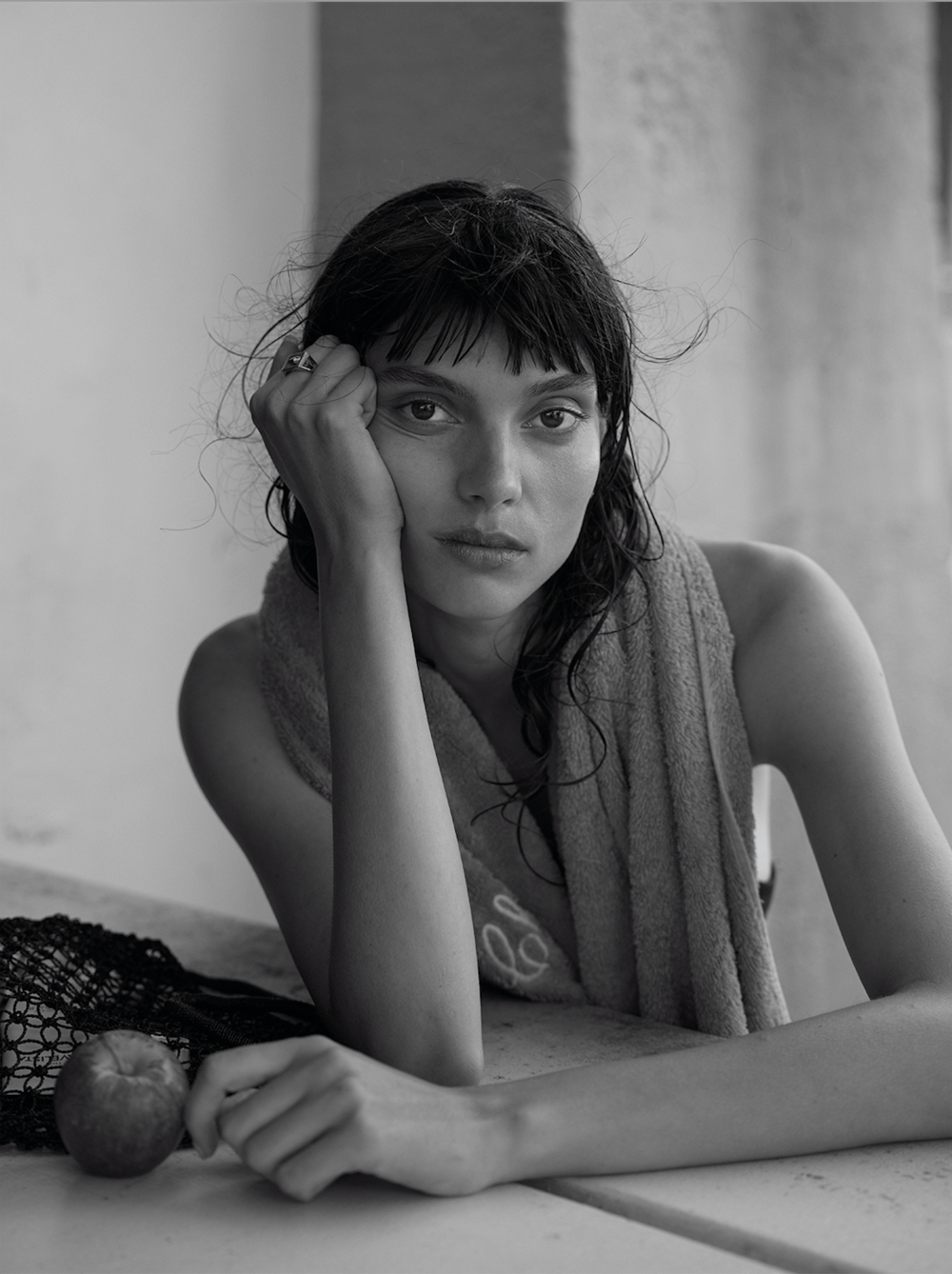 Monochrome portrait of a young adult woman with dark hair and a towel draped over her shoulders leaning on a table with an apple.