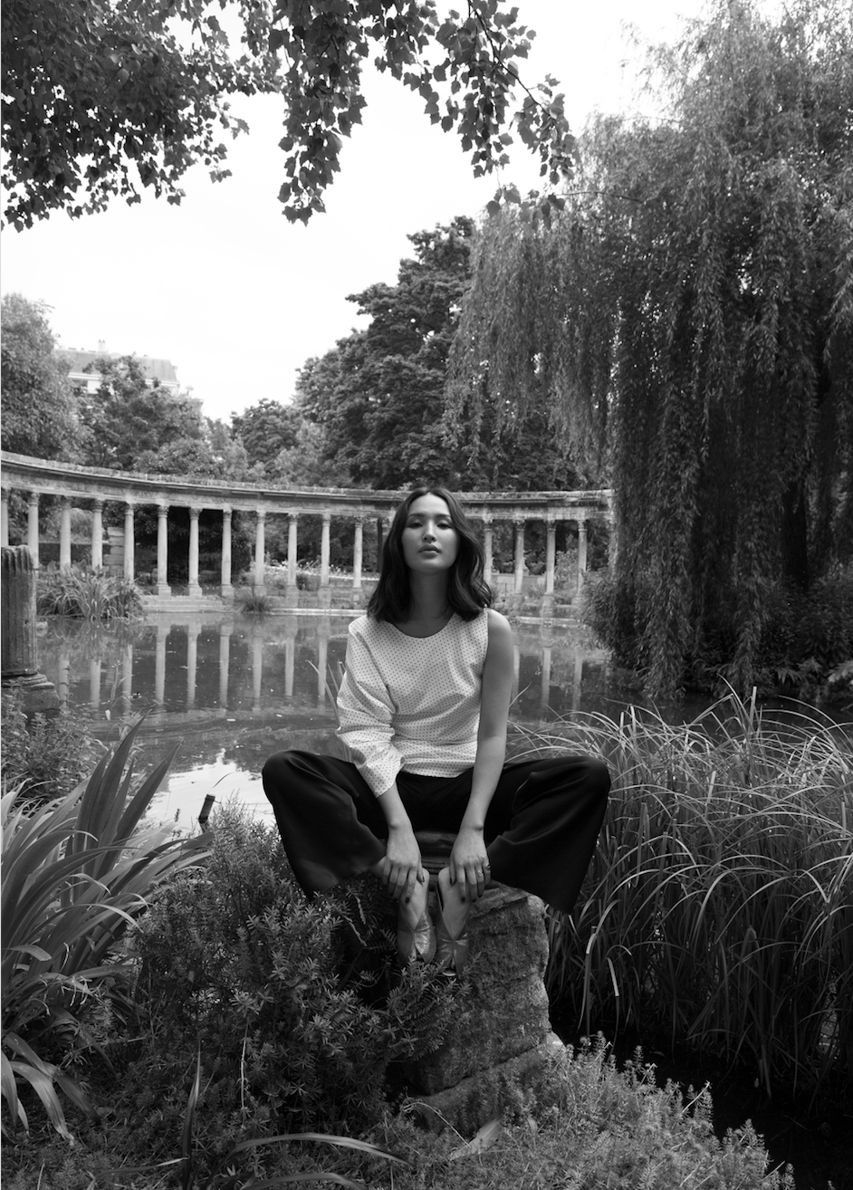 Black and white portrait of a woman sitting in a meditative pose in a park-like setting with classical architecture and lush greenery.