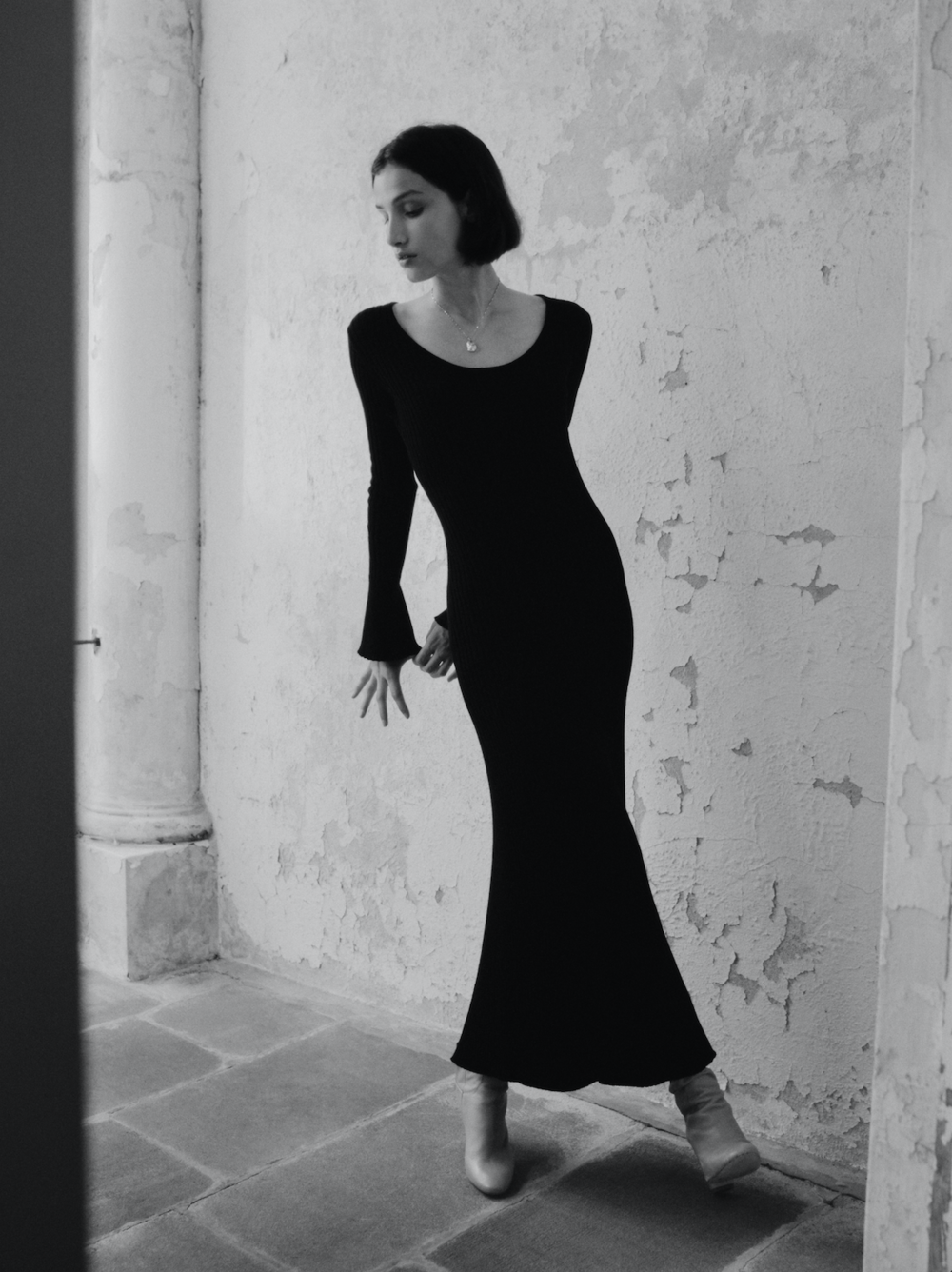A full shot of a woman in a long-sleeved black dress and boots, posing in an indoor setting with a stone floor and textured wall.