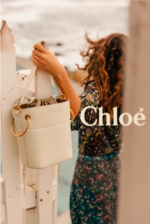 A woman holds a white Chloé bucket bag by a white fence near the ocean; the Chloé logo is visible in a fashion editorial.