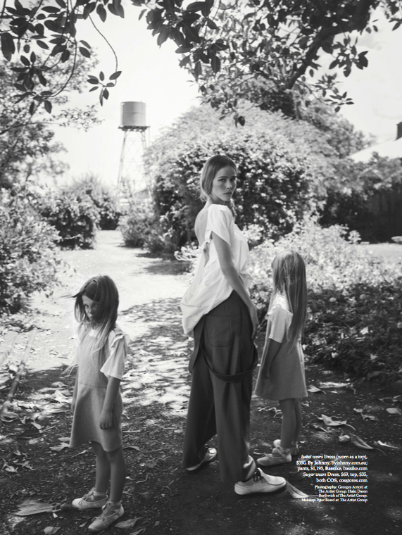 Black and white shot of an adult woman and two children standing outdoors in a garden setting near a water tower, wearing casual clothing.