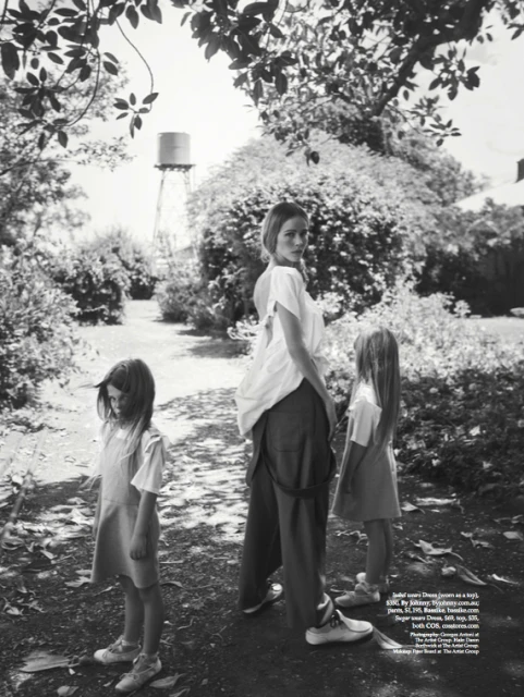 Black and white shot of an adult woman and two children standing outdoors in a garden setting near a water tower, wearing casual clothing.