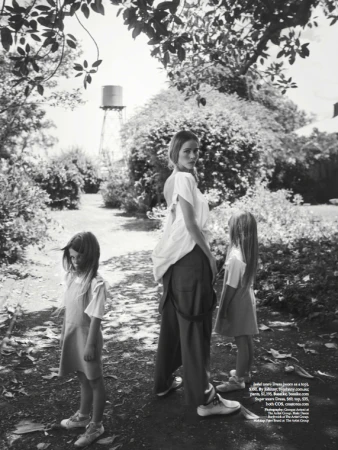 Black and white shot of an adult woman and two children standing outdoors in a garden setting near a water tower, wearing casual clothing.