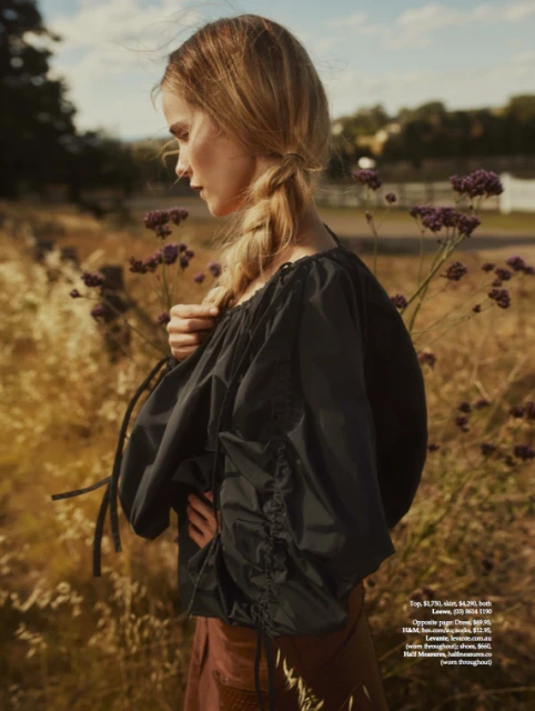 Woman in a black blouse stands in a field of tan grass and purple wildflowers with a blurred background in an outdoor fashion shot.
