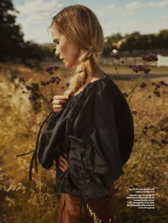 Woman in a black blouse stands in a field of tan grass and purple wildflowers with a blurred background in an outdoor fashion shot.